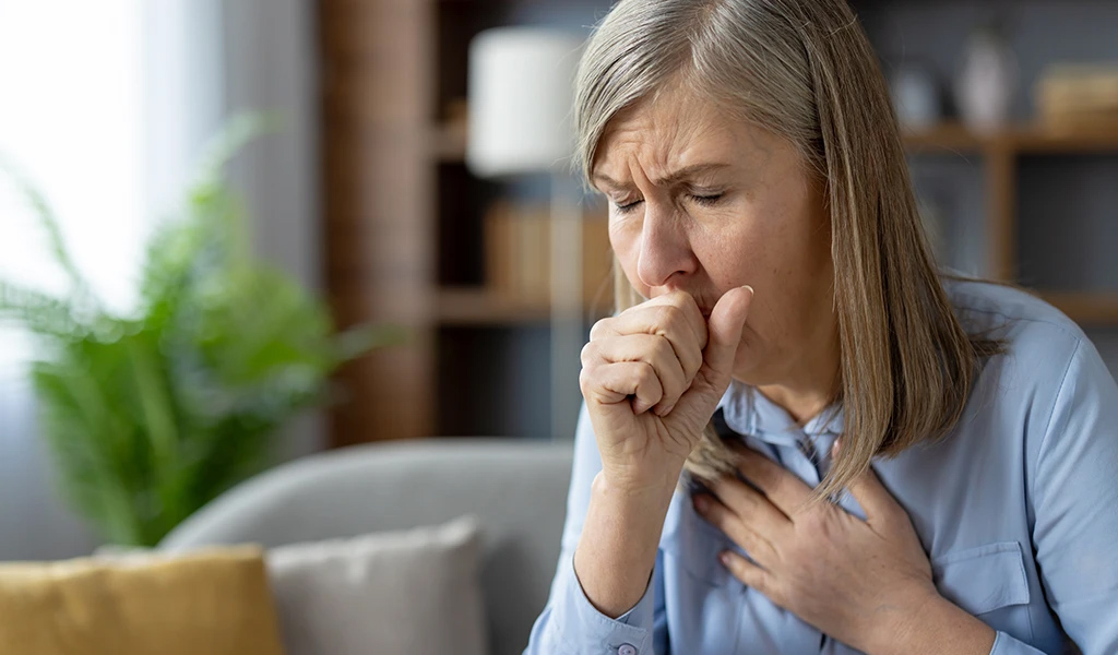 COPD Stufe 1 Ältere Frau, auf Sofa sitzend, hat einen Hustenanfall, und hält sich die Hand vor den Mund
