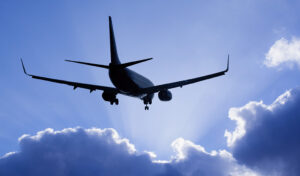 Ein Verkehrsflugzeug fliegt vor einem blauen Himmel mit Sonnenstrahlen und Wolken im Hintergrund.