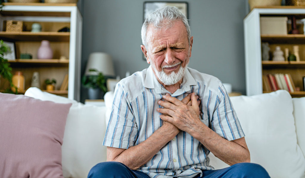 Person sitzt auf dem Sofa, hält beide Hände an die Brust und wirkt kurzatmig, Symbolbild für Atemnot und die Frage „Atemnot was tun?“