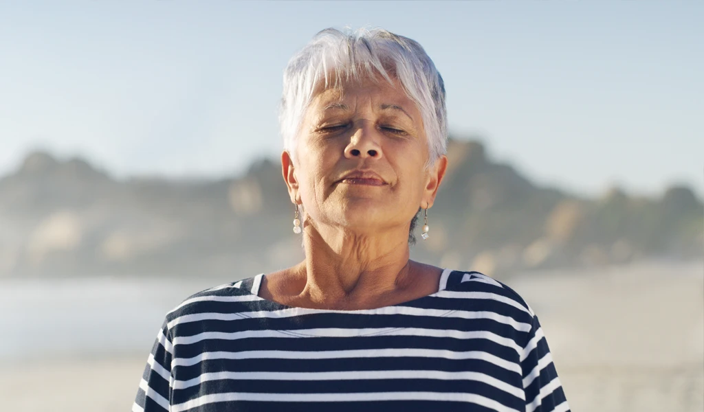 Salzinhalation bei Asthma und COPD Eine ältere Frau mit kurzen weißen Haaren und gestreiftem Hemd steht mit geschlossenen Augen an einem Strand.