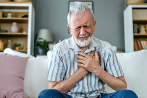 Person sitzt auf dem Sofa, hält beide Hände an die Brust und wirkt kurzatmig, Symbolbild für Atemnot und die Frage „Atemnot was tun?“