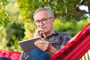 Ein älterer Mann mit Brille sitzt in einer Hängematte und hält ein Tablet in der Hand