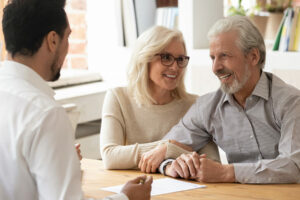 Lächelndes älteres Paar hält Händchen und unterhält sich mit einem Fachmann an einem Schreibtisch in einem hellen Büro.