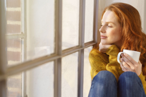 Frau mit roten Haaren in einem gelben Pullover hält eine Tasse und blickt friedlich aus einem sonnenbeschienenen Fenster.