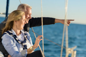 Lächelndes Paar auf einem Segelboot, das auf das Meer hinausblickt, während der Mann zum Horizont zeigt.
