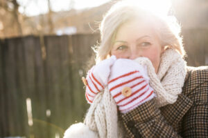 Lächelnde ältere Frau in Winterkleidung hält an einem sonnigen Tag draußen einen Schal vors Gesicht.