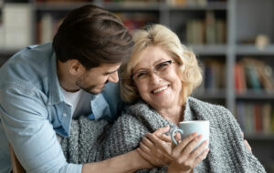Lächelnde ältere Frau mit einer Tasse, in eine Decke gehüllt, von einem jüngeren Mann in einem gemütlichen Raum umarmt.