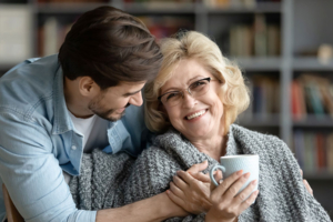 Lächelnde ältere Frau mit einer Tasse, in eine Decke gehüllt, von einem jüngeren Mann in einem gemütlichen Raum umarmt.