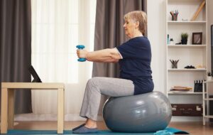 Eine ältere Frau sitzt auf einem Gymnastikball, stemmt Hanteln und schaut dabei in einem gemütlichen Wohnzimmer auf ein Tablet.