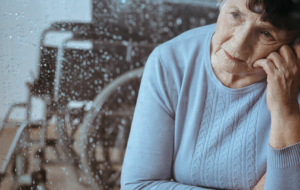 Traurig blickende ältere Frau am Fenster, im Hintergrund ein Rollstuhl.