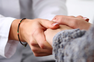 Nahaufnahme von zwei Personen, die sich an den Händen halten. Eine Person greift mit beiden Händen sanft die Hand der anderen, was einen Ausdruck von Mitgefühl, Unterstützung und Trost vermittelt.